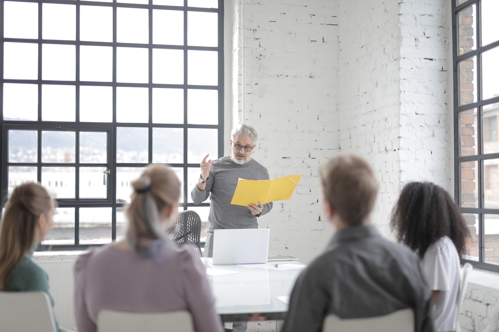 Serious middle aged executive in eyeglasses talking to diverse subordinates while taking yellow folder and standing in front of open laptop in light office